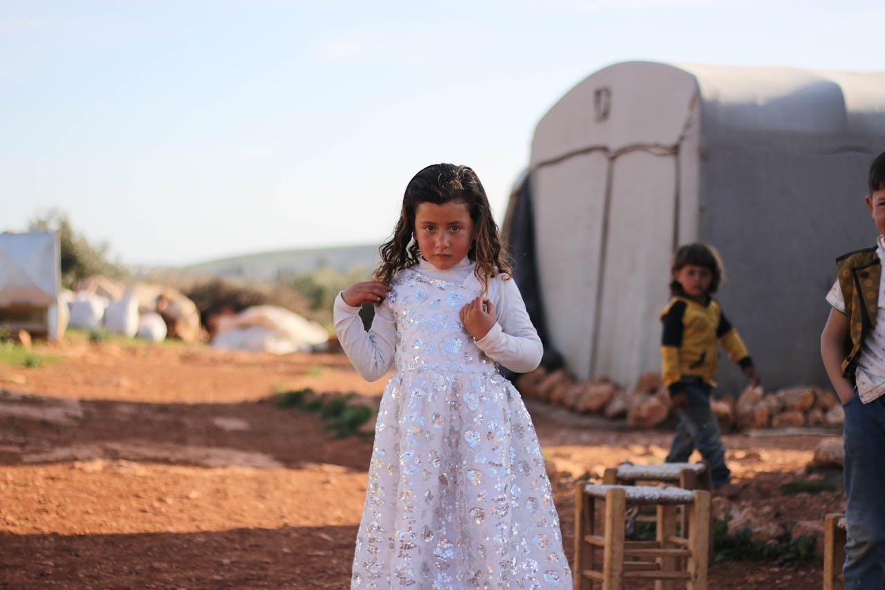 about-03 A young girl in a white dress stands in a refugee camp in Idlib, Syria, during summer.